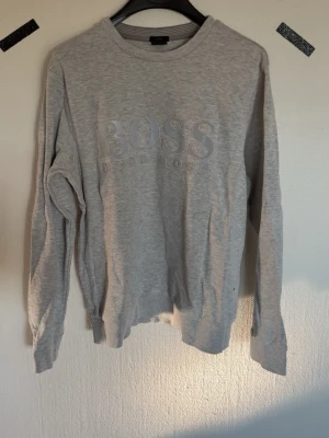 Grå sweatshirt från BOSS - Snygg grå sweatshirt från BOSS med diskret logga i relief på bröstet. Tröjan har rund halsringning, långa ärmar och ribbade muddar vid ärmslut och nederkant. Perfekt för en avslappnad och stilren look.