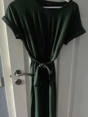 Mörkgrön klänning med knytband - Mörkgrön klänning från Twist & Tango med rund halsringning, korta ärmar och ett matchande knytband i midjan. Klänningen har en rak och enkel design som ger en avslappnad men ändå stilren look.