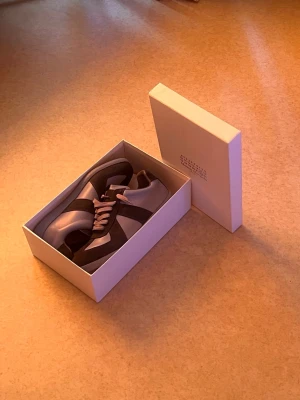 Maison Margiela  - Maison Margiela sneakers i grått och svart med vita detaljer. Klassisk låg modell med ovandel i läder och mocka, samt vita snören. Snygg minimalistisk design och levereras i originalkartong.