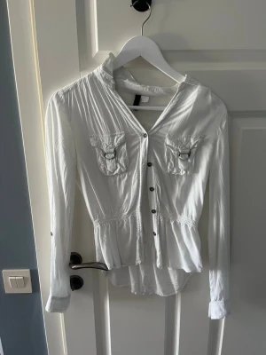 Vit blus med fickor och knappar - Snygg vit blus med långa ärmar, knappar framtill och två bröstfickor med lock. Blusen har markerad midja och lätt rynkade detaljer för en feminin siluett. Perfekt att styla med jeans eller kjol för en fräsch look.