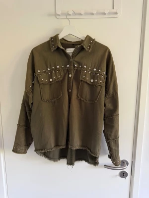 Zara vintage  - Cool olivgrön skjorta med råa kanter och nitar på bröstet, axlarna och ärmsluten. Skjortan har två bröstfickor och en avslappnad passform. Perfekt för dig som gillar en edgy och avslappnad stil.