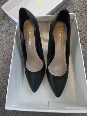Svarta pumps från Doria med spetsig tå - Stilrena svarta pumps från Doria med spetsig tå och hög klack. Skorna har en vågig kant längs öppningen och är tillverkade i ett slätt, matt material. Perfekta för dig som vill ha en klassisk och elegant look. Storlek 37