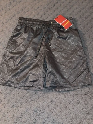 Svarta shorts GG shorts  - Snygga svarta shorts med diskret GG-logomönster över hela tyget. Shortsen har elastisk midja med snörning och är tillverkade i ett glansigt material som ger en lyxig känsla. Perfekta för dig som vill sticka ut med stil. PASSAR M. HELT NY, OANVÄND.