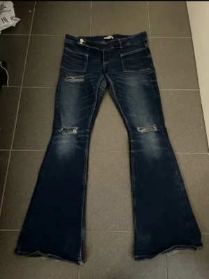 Mörkblå bootcut jeans med slitningar - Säljer ett par mörkblå bootcut jeans❤️dom är ifrån gina 14+ i storlek M, rätt så korta i storleken❣️ längst ner på byxorna är dom lite slitna eftersom jag gått ute med dom och då var dom för långa och har slitits i marken