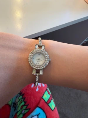 Silverfärgad klocka med strassdetaljer - Stilren klocka i silverfärg med rund urtavla och massor av glittrande strass runt både urtavlan och armbandet. Klockan har en smal länk och klassisk design som ger en lyxig känsla. Perfekt accessoar för att lyfta din outfit.