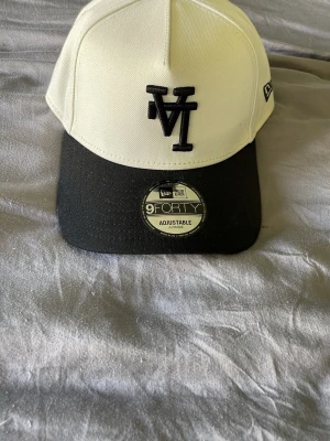 Vit och svart New Era 9FORTY keps - Snygg vit och svart keps från New Era, modell 9FORTY. Kepsen har justerbar passform, svart böjd skärm och broderad logga framtill samt New Era-logga på sidan. Tillverkad i slitstarkt tyg, perfekt för en cool och sportig look.