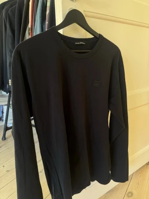 Svart långärmad T-shirt från Acne Studios - Svart långärmad topp från Acne Studios i 100% bomull. Enkel och stilren design med rund halsringning och diskret logotyp på bröstet. Perfekt basplagg för en clean och modern look.