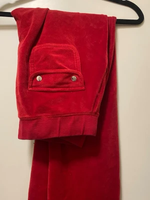 Röda velourmjukisbyxor Juicy Couture - Snygga röda mjukisbyxor i velour från Juicy Couture. Byxorna har en bakficka med två silvriga knappar och bred mudd vid benslut. Perfekta för en chill och trendig look.