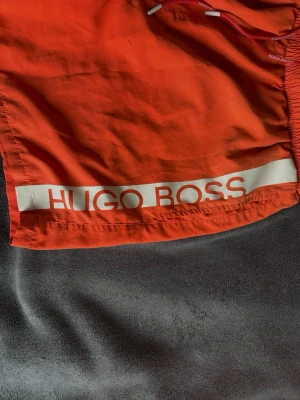 Orange badbyxor från Hugo Boss - Snygga orange badbyxor från Hugo Boss med vit logga längs ena benet. Klassisk modell med resår och dragsko i midjan. Perfekta för stranden eller poolen, tillverkade i snabbtorkande material.