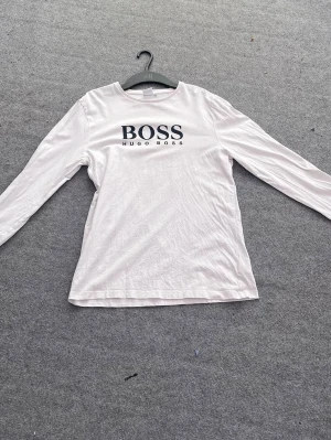 Vit långärmad topp från Hugo Boss - Snygg vit långärmad topp från Hugo Boss med stor logga framtill. Tillverkad i mjuk bomull som känns skön mot huden. Enkel och stilren design som passar till det mesta. Perfekt för dig som gillar klassiska plagg med en modern touch.