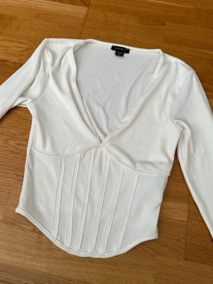 Vit långärmad korsettopp  - Snygg vit långärmad topp med korsett-inspirerad midja och djup v-ringning. Toppen har markerade sömmar framtill som ger en cool siluett och är gjord i mjukt material. Perfekt för dig som vill ha en trendig och stilren look.