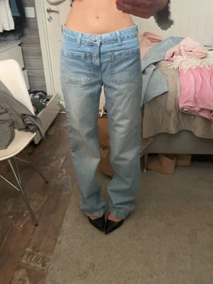 Ljusblå raka jeans med fickor - Säljer ett par ljusblå jeans med raka ben och stora framfickor. Jeansen har hög midja och klassisk knappgylf. Perfekta för en avslappnad och trendig look. Materialet är denim i bomull och passformen är loose fit.