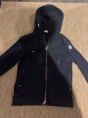Svart hoodie från Moncler med dragkedja - Snygg svart hoodie från Moncler med hel dragkedja framtill, två fickor med dragkedja och klassisk Moncler-logga på ärmen. Huva med dragsko och detaljer i rött och vitt på snöret. Perfekt för en stilren och sportig look.