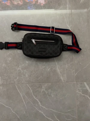 Gucci axelväska med monogram - Snygg svart axelväska från Gucci med klassiskt GG-monogram och justerbart band i rött och blått. Väskan har en silverfärgad dragkedja framtill och präglad logotyp. Perfekt för att bära det viktigaste med stil.