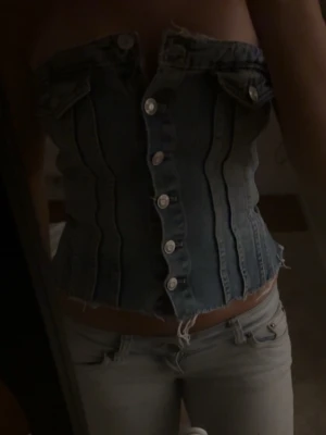 Blå denim korsett med knappar - Trendig blå denim korsett med knappar framtill och råa kanter. Toppen är axelbandslös och har dekorativa fickor upptill. Perfekt för dig som vill sticka ut med en edgy och cool stil.