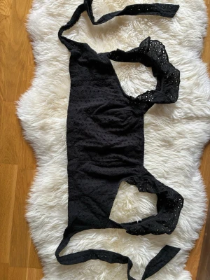 Svart broderad omlottopp med volang - Svart omlottopp med broderade detaljer och volang på axelbanden. Toppen knyts i midjan och har en feminin, ärmlös design. Perfekt för dig som vill ha en trendig och söt look med extra detaljer.