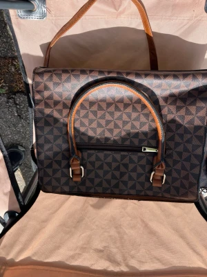 Brun handväska med geometriskt mönster - Snygg brun handväska med geometriskt mönster i mörkbrunt och ljusbrunt. Väskan har detaljer i ljusbrunt konstläder, dragkedja framtill och flera fack inuti. Perfekt för dig som gillar stilrena accessoarer med unik design.