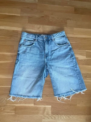 Ljusblå jeansshorts med rå kant - Säljer ett par ljusblå jeansshorts med klassisk femficksdesign och rå, fransig kant nedtill. Shortsen har en lite längre passform och är perfekta för sommaren. Har bälteshällor samt knapp- och dragkedjestängning. Använda några enstaka gånger 