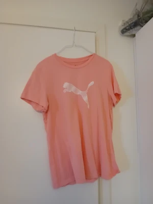 Rosa Puma t-shirt med logga - Säljer en rosa t-shirt från Puma med klassisk vit logga på bröstet. T-shirten har rund hals och korta ärmar, perfekt för dig som gillar sportiga och stilrena plagg. Passformen är normal och materialet känns mjukt och bekvämt.