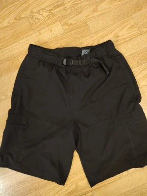Svarta cargoshorts med bälte herr stl S - Svarta cargoshorts med justerbart bälte i midjan och flera praktiska fickor. Shorts i en ledig passform, perfekta för en avslappnad stil. Materialet känns slitstarkt och passar bra till vardags. Enkel design utan synliga loggor. Bra skick. Herr stl S