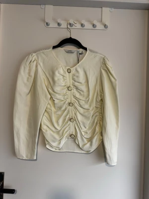 Krämvit blus från & Other Stories - Supersnygg krämvit blus från & Other Stories Paris Atelier med långa puffärmar och markerad rynkning framtill. Blusen har dekorativa knappar i guld och rund halsringning. Tillverkad i ett glansigt material som ger en lyxig känsla. Helt ny aldrig använd.