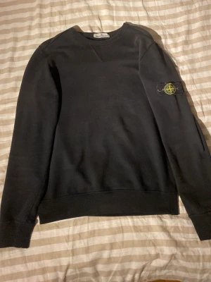 Svart sweatshirt från Stone Island - Svart sweatshirt från Stone Island med klassisk rund hals och ribbade muddar. Ikonisk logotyp-patch på vänster ärm. Perfekt för en stilren och avslappnad look.