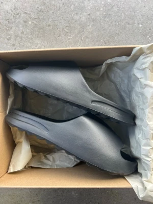 Svarta tofflor, Yeezy slide box. - Svarta tofflor med Yeezy Slide box. Har dock ett litet märke på vänster tofflan (bild 6).