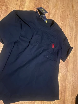 Svart t-shirt från Polo Ralph Lauren - Svart t-shirt från Polo Ralph Lauren med klassisk passform och korta ärmar. T-shirten har en röd broderad logga på bröstet och är gjord i mjuk bomull. Perfekt för en stilren och enkel look.