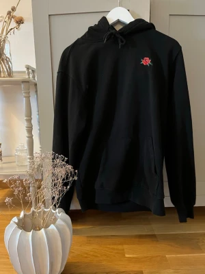 Svart hoodie med broderad ros - Cool svart hoodie med huva och en liten broderad röd ros på bröstet. Tröjan har en klassisk känguruficka och dragsko i huvan. Perfekt för dig som gillar stilrena plagg med en twist. Materialet känns mjukt och bekvämt.