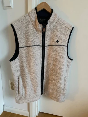 Morris väst - Mysig beige teddyväst från Morris med svart kantband och dragkedja framtill. Västen har hög krage, två fickor och en liten broderad logga på bröstet. Perfekt för lager-på-lager och chill still. (Pris går att diskutera) !?