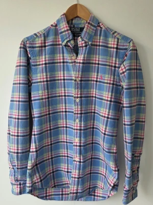 Rutig skjorta från Polo Ralph Lauren, stl s - Snygg rutig skjorta i blått, rosa, vitt och grönt från Polo Ralph Lauren. Skjortan har klassisk krage, knappar framtill och lång ärm. Tillverkad i mjuk bomull och har en broderad logga på bröstet.