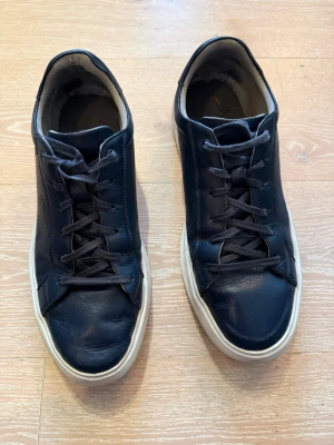 Mörkblå lädersneakers med vit sula - Stilrena mörkblå sneakers i läder med klassisk vit sula och svarta skosnören. Skorna har en låg profil och enkel design som passar till det mesta. Insidan är fodrad med ljus textil för extra komfort. Ett skosnöre behöver bytas