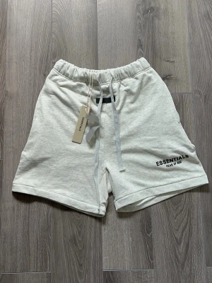 Essentials Fear of God shorts ljusgrå - Snygga ljusgrå shorts från Essentials Fear of God med elastisk midja och snörning. Mjukt bomullsmaterial och logga tryckt på vänster ben. Perfekta för en chill och avslappnad stil.