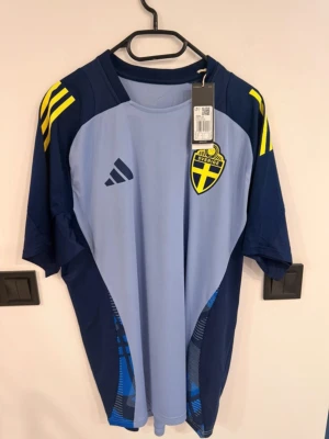 Svenska landslagets träningströja Adidas - Snygg ljusblå och marinblå träningströja från Adidas med Sveriges landslagsemblem på bröstet. Tröjan har gula detaljer på axlarna och är tillverkad i lätt och ventilerande AEROREADY-material. Perfekt för fotbollsträning eller sportiga aktiviteter.
