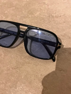 Svarta Tom Ford pilotbågar solglasögon - Snygga svarta solglasögon från Tom Ford med klassisk pilotbåge och blåtonade glas. Ramen är i blank svart plast med gulddetalj vid gångjärnet. Perfekt accessoar för en cool look.