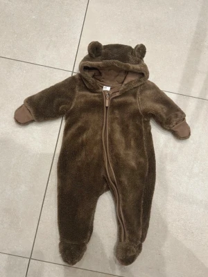Brun teddyoverall med öron - Supersöt brun teddyoverall med huva och små björnöron. Dragkedja framtill och mjukt, fluffigt material som känns riktigt gosigt. Overallen har vikbara muddar vid ärmar och ben för extra värme. Perfekt för kyliga dagar!   Trikåfodrad!   Obs! Stygnen har sprätts upp lite vid halsen, går lätt att sy. Se bild