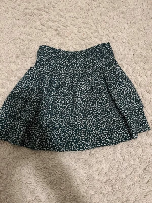 Blommig kortkjol från Zara - Supersöt kortkjol från Zara med blommigt mönster i mörkgrönt och vitt. Kjolen har en smockad midja som ger en bekväm passform och ett lätt utsvängt fall. Perfekt för dig som gillar en lekfull och trendig stil.