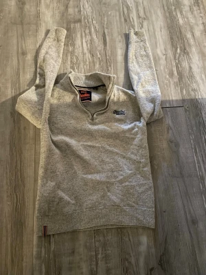 Grå halvzip-tröja från Superdry - Snygg grå långärmad tröja från Superdry med halvzip och broderad logga på bröstet. Tröjan har ribbade muddar och krage, perfekt för lager-på-lager. Mjuk och skön känsla, passar till jeans eller chinos.