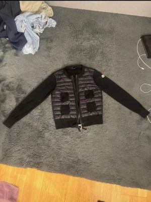 Svart quiltad pufferjacka Moncler - Säljer en svart pufferjacka från Moncler i storlek S. Jackan har quiltad front i blankt material, stickade ärmar och ribbade muddar. Fyra fickor framtill och dragkedja hela vägen. Klassisk Moncler-logga på ena ärmen. Har ett litet hål på baksidan vid nacken men inget som syns. Skriv gärna dm ifall du skulle vilja ha mer bilder på cardiganen eller frågor.