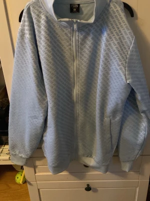 Blå hoodie med dragkedja och tryck - Säljer en grå hoodie från CHOICE med hel dragkedja framtill och ett mönster av upprepat logotyp-tryck över hela plagget. Hoodien har huva med dragsko och ribbade muddar vid ärmslut och nederkant. Perfekt för en chill och avslappnad stil.