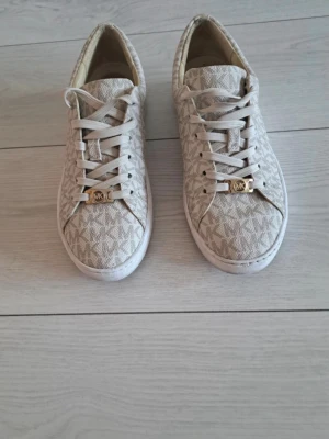 Beige MK sneakers med monogram - Snygga beige sneakers från Michael Kors med klassiskt MK-monogram över hela skon. Skorna har vita snören och guldfärgade detaljer framtill. Tillverkade i textil med gummisula och låg profil för en clean look. 