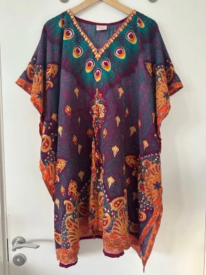 Färgglad kaftan med påfågelfjädrar - Snygg strandkaftan i tunnt, flowy material med färgstarkt mönster av påfågelfjädrar och paisley i lila, orange och grönt. V-ringad hals med dekorativa detaljer. Perfekt att slänga över bikinin på stranden. One-size, justerbart midjeband, från XS till XL. 