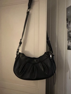 Svart axelväska i skinnimitation - Snygg svart axelväska i skinnimitation med justerbart axelband och silverfärgade detaljer. Väskan har en rundad form och dekorativa sömmar framtill. Perfekt storlek för det viktigaste och ger en cool vibe till din outfit.