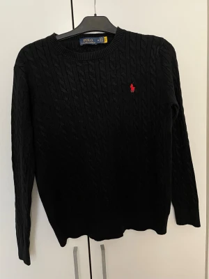 Svart kabelstickad tröja Polo Ralph Lauren - Kabelstickad svart tröja från Polo Ralph Lauren med rund halsringning och röd broderad logga på bröstet. Tröjan har långa ärmar och är tillverkad i mjukt material som passar perfekt till lager-på-lager-stil.