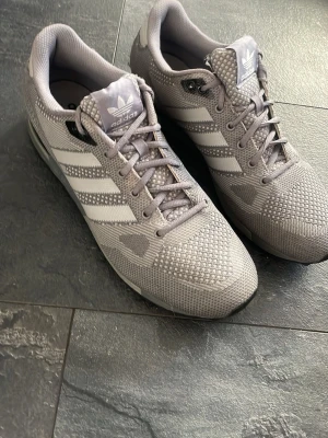 Grå Adidas ZX 750 sneakers - Snygga grå Adidas ZX 750 sneakers med klassiska tre ränder på sidorna och meshdetaljer för extra andningsförmåga. Skorna har en sportig vibe, snörning och diskreta loggor på plös och häl. Perfekta för dig som gillar stilrena och bekväma sneakers. Dom är stora i storlek så skon är som 44 ungefär 