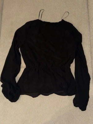 Svart omlottblus från Vila - Stilren svart omlottblus från Vila med långa ärmar och markerad midja. Blusen har tunna axelband och transparenta detaljer samt spets vid axlarna. Perfekt för en trendig och elegant look.