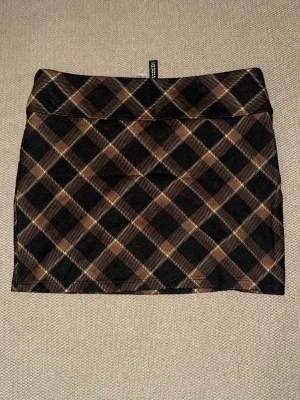 Rutig brun och svart minikjol - Snygg kort kjol  i rutigt mönster med bruna, svarta och beige toner. Kjolen har en rak passform och är perfekt för att skapa en trendig look. Tillverkad i mjukt och stretchigt material för extra komfort.