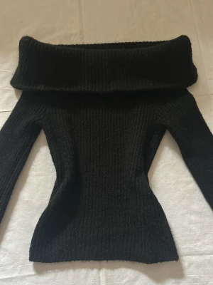 Svart off shoulder stickad tröja - Trendig svart stickad tröja med off shoulder-design och bred krage. Tröjan är ribbstickad och har lång ärm, perfekt för en snygg och bekväm look. Passar till många olika outfits och är ett måste i garderoben.