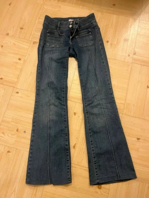 Mörkblå bootcut jeans med hög midja - Säljer ett par mörkblå bootcut jeans med låg midja och dubbla knappar framtill. Jeansen har fram- och bakfickor samt markerade sömmar längs benen för en snygg retrostil. Perfekta för dig som gillar 00-talsvibbar och vill ha en bekväm passform.