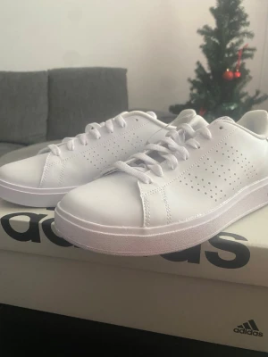 Vita Adidas sneakers med rosa logga - Fräscha vita sneakers från Adidas med klassisk låg profil och perforerade detaljer på sidorna. Skorna har vit sula, vita skosnören och rosa Adidas-logga på hälen. Tillverkade i syntetmaterial med en stilren och clean look.